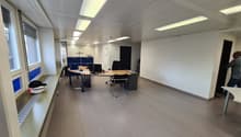 The image shows a modern, open-plan office space with a tiled floor, suspended ceiling with recessed lighting, and several desks with computers and office chairs. The space appears to be well-lit and organized, with a Proman sign visible in the corner.