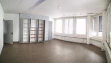 Large open room with white walls, hardwood flooring, and multiple built-in shelving units along the walls. The room has several large windows providing natural light.