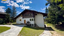 2-story chalet-style house with wooden beams, white walls, and large windows. The house is surrounded by a lush green lawn and pine trees, with a stone pathway leading to the entrance. There appears to be a balcony or terrace on the upper level.