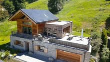 2-story wooden and stone house with a slanted roof, large balcony, garage, and parking space. The house is surrounded by a lush green landscape with trees and vegetation.