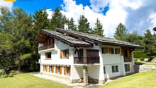 Alpine-style house, white exterior, wooden balcony, green roof, large windows, surrounded by a green lawn and trees