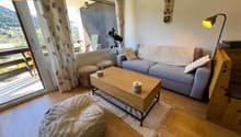 Cozy living room with large windows, gray sofa, wooden coffee table, and a telescope on a tripod. The room has a balcony or terrace with a scenic mountain view.