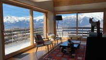 large glass windows, wooden floor, carpet, glass door to balcony, coffee table, chairs, statue, rug, mountains view
