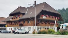 A multi-story wooden building with a tiled roof, balconies, and flower boxes. There are cars parked in front of the building.