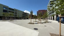 The image shows a modern residential area with multi-story apartment buildings. The buildings have balconies and are surrounded by a paved plaza with trees and benches. The area appears to be well-designed and maintained, with a clean and open layout.