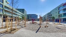 The image shows a public plaza or outdoor space in an urban setting. The area is paved with gravel and features various wooden structures, possibly seating or play equipment. The surrounding buildings have balconies and appear to be modern, multi-story re