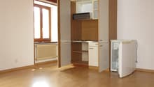 Empty room with wooden floor, white walls, kitchen area with open cabinets, window, and a refrigerator.