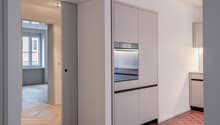 Minimalist modern kitchen, white cabinets, wooden floor, red tiles near oven, sliding door leading to another room, open window in other room.