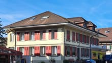 Building has brown tiled roof, yellow walls, and red shutters on windows, garden area with plants, bench, and a car parked nearby