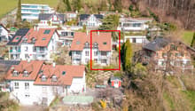 Aerial view of a residential area with several houses, one of which is highlighted with a red square. The highlighted house has a brown roof and white walls. The houses are surrounded by trees and greenery. The area is on a slope, with some houses having