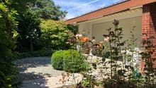Garden with gravel path, various plants and flowers, brick building with garage, and a white chair.