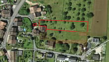 This image shows an aerial view of a residential area with several buildings and a large open green space. The highlighted area appears to be an undeveloped plot of land, possibly a vacant lot or agricultural field. There are some parking spaces visible n