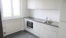 Modern kitchen with white cabinets, stainless steel countertop, built-in oven, and a sink with a faucet. It has a window with a white frame. The floor is tiled with gray tiles.