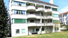 3-story apartment building with white exterior, balconies on each floor, and large windows. The building has a green roof and is surrounded by a grassy area with trees.