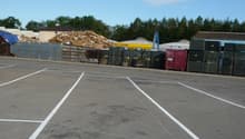 Paved parking lot with marked parking spaces, a large pile of wood debris, and various storage containers or dumpsters in the background.