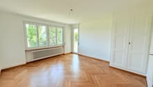 Spacious room with large windows, hardwood parquet flooring, and a radiator. The room appears to be empty, with white walls and a door leading to another area.