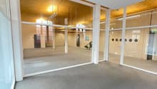 The image shows a large, open commercial space with a wooden ceiling and walls. The space has a grey carpeted floor and large glass windows that allow natural light to enter. There are various lighting fixtures and equipment visible, suggesting this could
