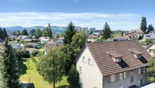 The image shows a residential neighborhood with a mix of single-family homes and multi-story apartment buildings. The area is surrounded by lush greenery, including trees and well-maintained lawns. The buildings have a mix of architectural styles, with so