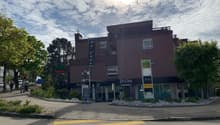 A multi-story commercial building with a Migros grocery store and other businesses on the ground floor. The building has a modern, dark-colored exterior with large windows. There are trees and landscaping around the building, and a crosswalk in front of t