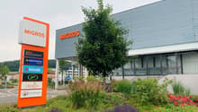 Large Migros supermarket building with several service stores, tree, and well-maintained flower beds in front