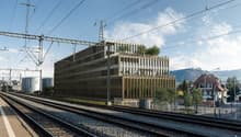 Modern office building with glass facade, located near a railway track, surrounded by trees and a house