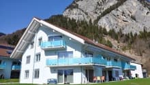 A multi-story apartment building with balconies, surrounded by a grassy area and with mountains in the background.
