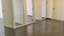 The image shows an empty office space with a tiled floor and glass-paneled doors. The room has a clean, modern appearance with neutral-colored walls and overhead lighting.