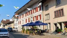 This image shows a street view of a quaint European town. The buildings have traditional architecture with red shutters and wooden beams. There are several restaurants and shops along the street, with outdoor seating areas under blue umbrellas. Bicycles a
