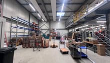 Spacious warehouse with high ceilings, multiple shelves and storage racks, various boxes and materials, trolleys and pallets, stairs, and metal beams.