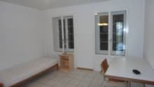 small room with single bed, wooden table, desk, wooden chair, lamp on table, white walls, windows, white tile floor