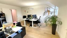 Spacious office space with hardwood floors, white walls, adjustable desks, computers, and a large potted plant. The room appears to have a balcony or terrace visible through the window.