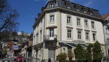 Corner building, 3 stories, white facade, multiple windows, beige trims, black roof, balcony with railings, awnings, and a name 'LANDHAUS' on the facade
