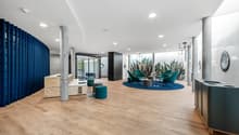 Spacious lobby with hardwood floors, white walls, blue curtains, modern furniture including green armchairs and ottomans, large wall mural with tropical plants, and various storage units.