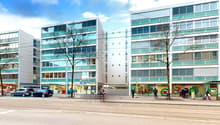 Three buildings with large windows, on a street, with people walking, cars parked, trees, and shops in front.