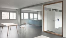 Spacious open-plan living area with a kitchen, dining table, and living room. Large windows provide ample natural light. The kitchen features blue cabinets and a minimalist design. The dining table and chairs are made of wood, complementing the overall mo