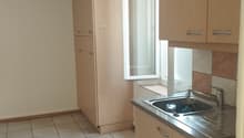 The image shows a kitchen area with beige cabinets, a sink, and a refrigerator. The floor is tiled, and there is a window visible. The kitchen appears to be part of a larger living space.