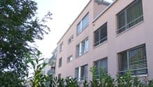 Multiple story building, pink paint, multiple windows with blinds, balcony, greenery
