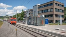 A train station on a sunny day. The station has a large brown building, two sets of train tracks, a shelter with a bench, and a red train approaching.