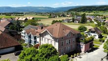 The image shows a scenic rural town with a mix of residential and commercial buildings. The buildings have red tile roofs and are surrounded by lush greenery, including trees and fields. There are several multi-story apartment buildings with balconies vis
