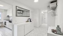 Modern white kitchen with ceramic tiles, white cabinets, kitchen counter, gas stove, sink, dishwasher, ceramic baskets, basket on the wall, hanging rack, lamp, mirror, table with a gray vase and a plate