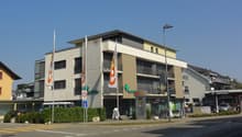 multi-story building with ground floor, pharmacy on ground floor, cars parked in front, greenery, streetlights, bicycles, bicycles