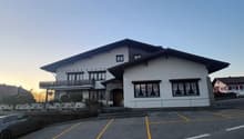 Two-story building with a traditional Bavarian-style architecture, featuring a sloped roof and white exterior walls. The building has several windows and a balcony on the upper level. There is a parking lot in front of the building.