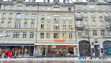 A multistory building with multiple entrances to stores, people walking in front, bicycles parked, trees, and a snowy ground