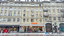 A multistory building with multiple entrances to stores, people walking in front, bicycles parked, trees, and a snowy ground