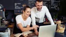 The image shows a couple working together at a cafe or restaurant. They are standing at a counter, with a laptop computer and a notebook in front of them. The background features a chalkboard menu and other cafe decor. The couple appear to be engaged in a
