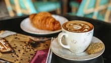 A cup of coffee with a latte art design, served with a croissant on a plate. The setting includes a newspaper and other items on the table, suggesting a cozy cafe environment.