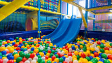 Colorful indoor playground with a blue slide, yellow climbing structures, and a ball pit filled with multicolored plastic balls.