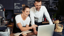 The image shows a couple working together at a cafe or restaurant. They are sitting at a counter, with a laptop and a book open in front of them. The background includes a chalkboard menu and other cafe equipment, suggesting this is a commercial food serv