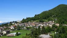 The image shows a picturesque mountain village nestled in a lush, green valley. The village is built on a raised ground, with houses and buildings scattered across the hillside. The landscape is dominated by the surrounding mountains, which are covered in