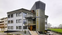 Contemporary building with multiple floors, modern facade, staircases leading to the entrance, car parked in front, and a surrounding landscape.